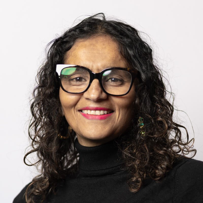Head and shoulder portrait of a smiling, middle-aged woman with glasses and wavy brown hair wearing a black roll neck top.